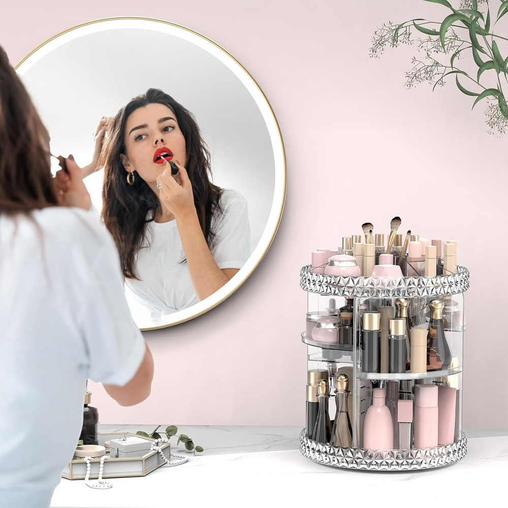 Woman applying makeup in front of a mirror with a clear cylindrical makeup organizer filled with products on a pink wall background.